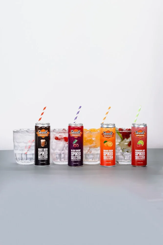 Four Stewart’s Spiked Seltzer cans with filled glasses on a pale studio background